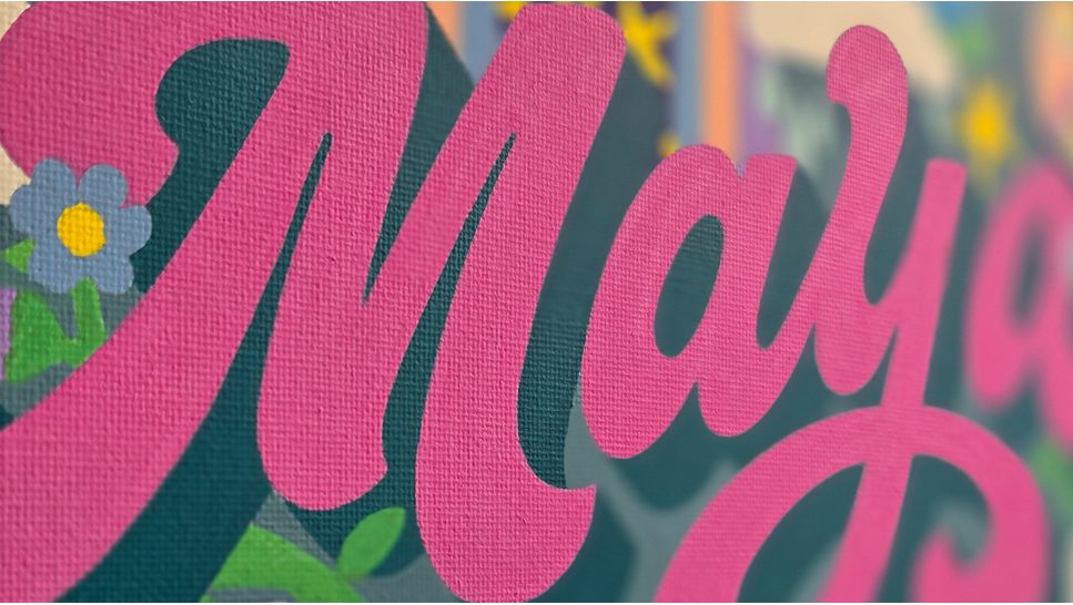 The name Maya hand-lettered in pink on a painted canvas with small flowers